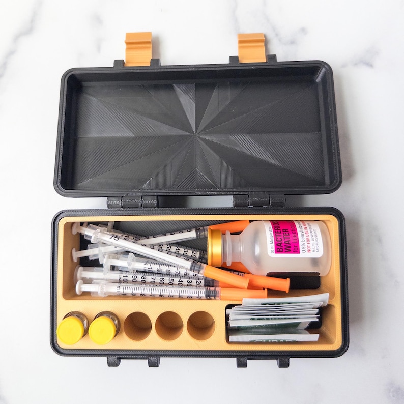 May include: Black and orange medical supply case, open to reveal syringes, vials, and other medical supplies. The case has a textured lid and a yellow interior. The case is on a white marble surface.