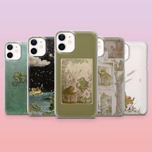 May include: A collection of five clear smartphone cases featuring various illustrations of two anthropomorphic frogs. The designs include scenes of frogs on a bicycle, in a boat under a starry sky, in a garden, and a tarot card style graphic.