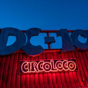 Puede incluir: Un edificio rojo con un gran letrero que dice "DC-10" en letras plateadas. Debajo del letrero hay un letrero de neón que dice "CIRCOLOCO" en letras rojas.