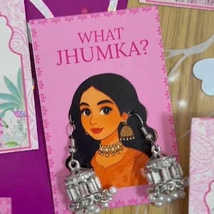 Może przedstawiać: R&oacute;żowa karta z napisem "WHAT JHUMKA?" przedstawia ilustrację kobiety noszącej srebrne kolczyki. Kolczyki są szczeg&oacute;łowe z przezroczystymi kamieniami i małymi białymi perłami. Kobieta na ilustracji ma czarne włosy i nosi pomarańczowy top.