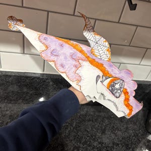 May include: A handmade dragon mask crafted from white paper, featuring a detailed design with purple, orange, and black ink. The mask has a whimsical, cartoonish style, with a spiderweb design on the horns and a star in the eye.