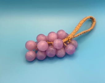 May include: A cluster of light purple, round beads resembling grapes, connected by a brown rope handle. The beads have a matte finish and are arranged in a bunch. The background is a solid light blue.