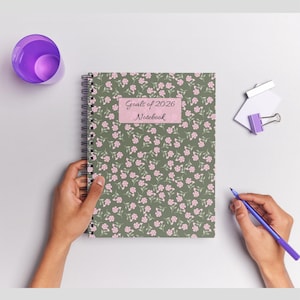 Op de afbeelding: Een spiraalgebonden notitieboek met een bloemenpatroon van roze bloemen op een groene achtergrond. De kaft heeft een roze label met de tekst "Goals of 2026 Notebook". Een paarse pen en beker liggen op het witte oppervlak.