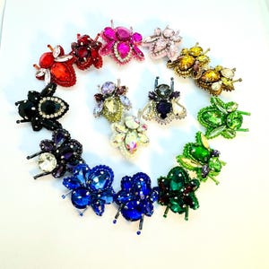 Könnte beinhalten: Eine Sammlung von 12 bunten Bienenbroschen, jede mit einem anderen Farbschema und Kristallverzierungen. Die Broschen sind in einem Kreis auf weißem Hintergrund angeordnet.