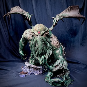 Può includere: Scultura dettagliata di una creatura mostruosa con grandi ali, tentacoli e una testa bulbosa. La figura è principalmente verde con sfumature marroni e una gemma rossa sulla testa. Si erge su una base che raffigura onde e una piccola barca.