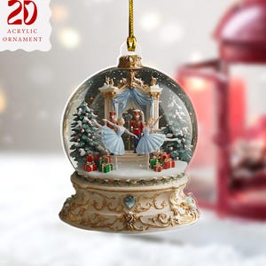May include: A 2D acrylic Christmas ornament in the shape of a snow globe. It features a stage with ballerinas, a nutcracker, and Christmas trees. The base is ornate, and it has a gold hanging string. The text on the ornament reads "2D Acrylic Ornament."