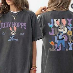 Peut inclure: T-shirt gris fonc&eacute; avec Judy Hopps de Zootopia. Le devant du t-shirt porte l'inscription "JUDY HOPPS" au-dessus d'une image de Judy Hopps, avec "Est 2025" et "Zootopia" en dessous. Le dos du t-shirt pr&eacute;sente une image plus grande de Judy Hopps.