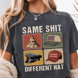 May include: Dark grey t-shirt featuring the text "SAME SHIT DIFFERENT HAT" and four hat illustrations. The hats depicted are a KKK hood, a Confederate flag hat, a military hat, and a "Make America Great Again" hat.