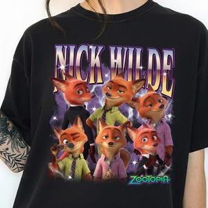 Puede incluir: Camiseta negra con un dise&ntilde;o de Nick Wilde de Zootopia. El dise&ntilde;o incluye el texto "NICK WILDE" en fuente morada y dorada sobre un grupo de zorros de dibujos animados. La palabra "Zootopia" est&aacute; en verde en la parte inferior.