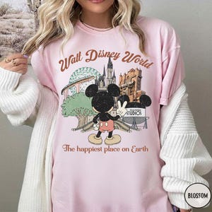 May include: A light pink t-shirt featuring a graphic of Mickey Mouse in front of Walt Disney World landmarks. The text "Walt Disney World" and "The happiest place on Earth" are printed in brown.