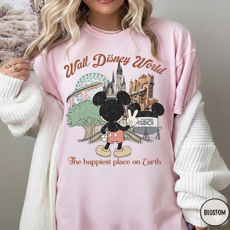 May include: A light pink t-shirt featuring a graphic of Mickey Mouse in front of Walt Disney World landmarks. The text "Walt Disney World" and "The happiest place on Earth" are printed in brown.
