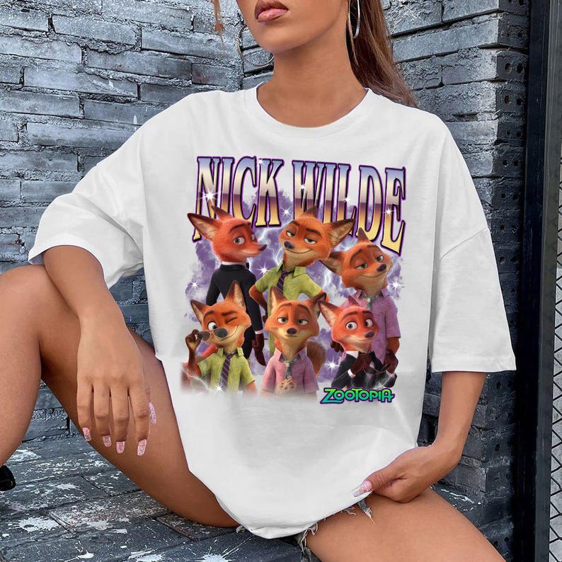 Puede incluir: Camiseta blanca con un dise&ntilde;o de Nick Wilde de Zootopia. El dise&ntilde;o incluye el texto "NICK WILDE" en fuente morada y dorada sobre un grupo de zorros de dibujos animados. La palabra "Zootopia" est&aacute; en verde debajo.