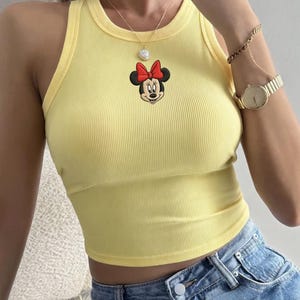May include: A yellow ribbed tank top featuring an embroidered Minnie Mouse design with a red bow. The top is paired with blue denim jeans. A gold watch and jewelry complete the look.