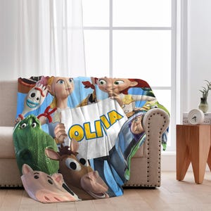 May include: A plush blanket featuring characters from the animated film Toy Story. The blanket displays a variety of characters, including Jessie, Buzz Lightyear, and others, with the name "OLIVA" prominently displayed. The blanket is draped over a beige sofa.