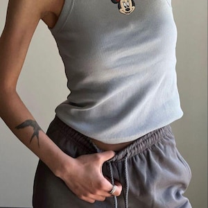May include: A light blue ribbed tank top featuring a Minnie Mouse pendant necklace with a red bow. The top is paired with gray sweatpants. The outfit is casual and comfortable.
