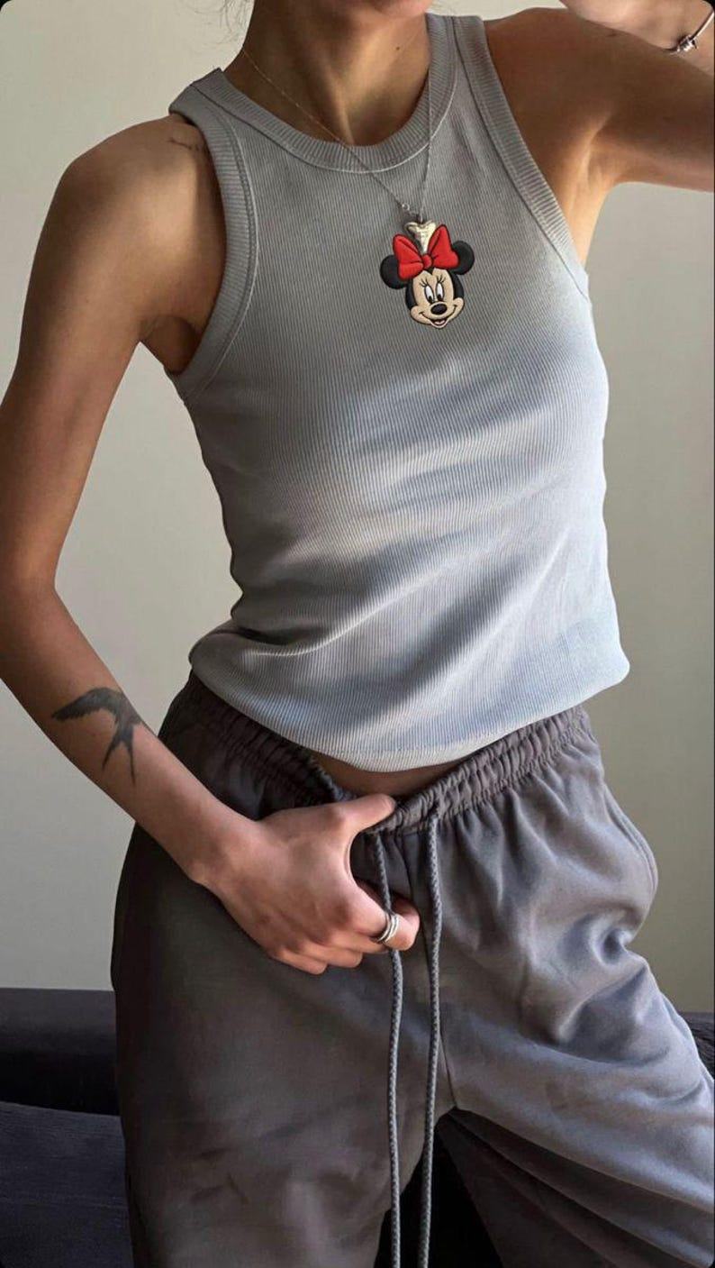 May include: A light blue ribbed tank top featuring a Minnie Mouse pendant necklace with a red bow. The top is paired with gray sweatpants. The outfit is casual and comfortable.