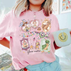 May include: Pink t-shirt featuring a colorful graphic of Toy Story characters in framed portraits, with the text "It's my birthday" and "Your name". The number "4" is also displayed. A white mug with a smiley face is held in the image.