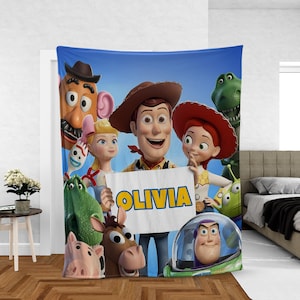 May include: A blue blanket featuring characters from the animated film Toy Story. The blanket displays Woody, Buzz Lightyear, Jessie, and other characters. The name "OLIVIA" is written on a sign held by Woody.