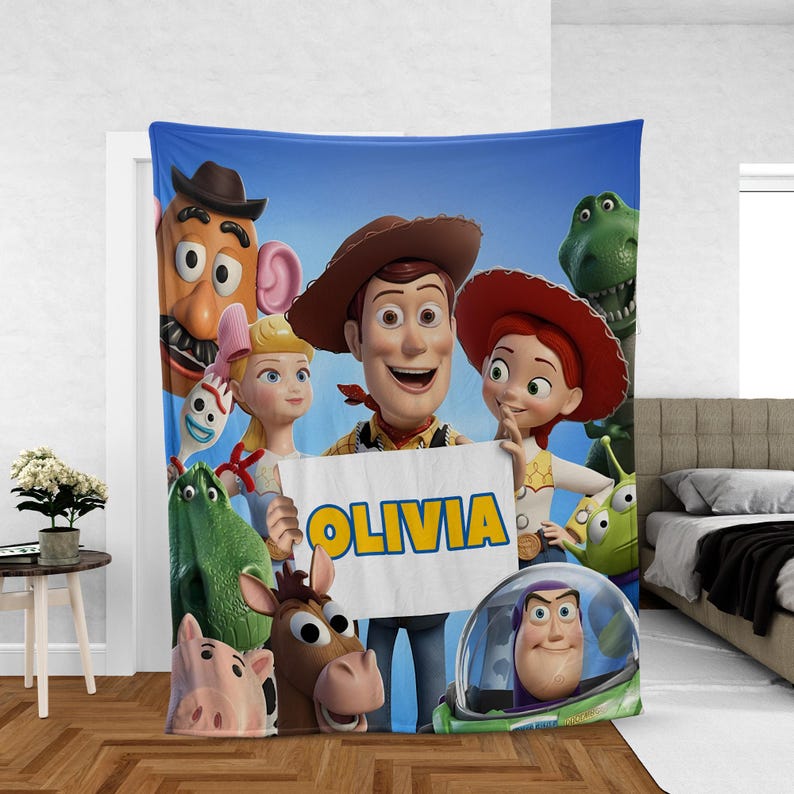 May include: A blue blanket featuring characters from the animated film Toy Story. The blanket displays Woody, Buzz Lightyear, Jessie, and other characters. The name "OLIVIA" is written on a sign held by Woody.