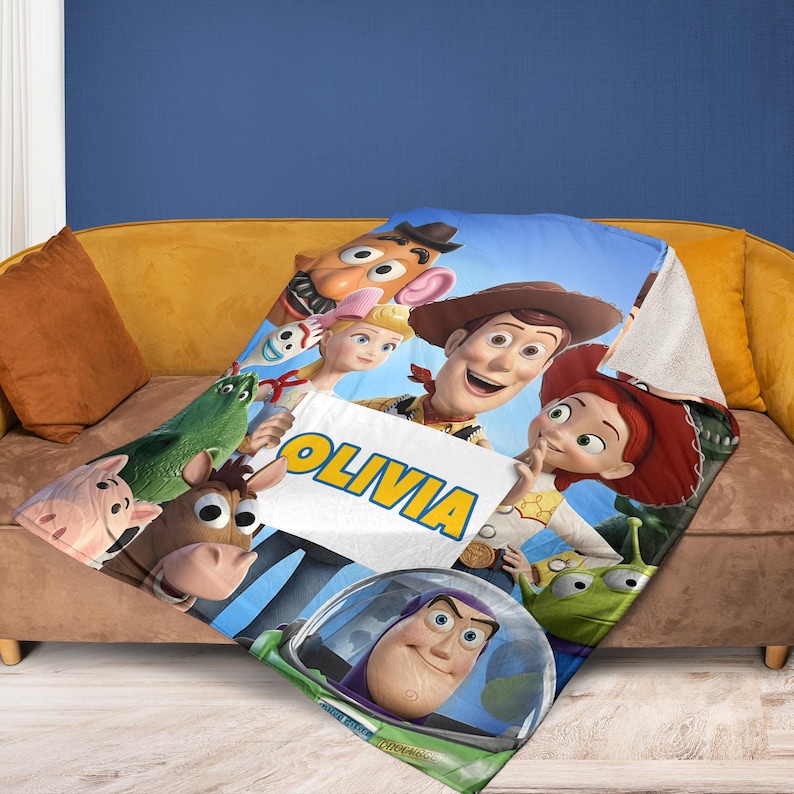 May include: A personalized blanket featuring characters from the animated film Toy Story. The blanket displays a blue sky background with Woody, Buzz Lightyear, Jessie, and other characters. The name "OLIVIA" is prominently displayed on a white banner.