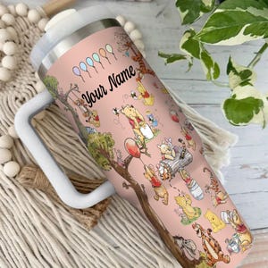 May include: A pink insulated tumbler featuring a whimsical design of Winnie the Pooh and friends. The tumbler has a white handle and lid, with the text "Your Name" in black script. The design includes balloons and various characters.