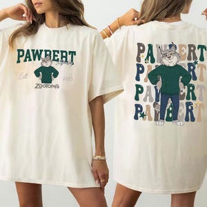 Peut inclure: T-shirt cr&egrave;me avec un personnage de dessin anim&eacute; de Zootopie. Le devant affiche le texte "PAWBERT" au-dessus du personnage, avec "Est 2025" et "ZOOTOPIA" en dessous. Le dos r&eacute;p&egrave;te "PAWBERT" avec le personnage au centre.