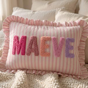 May include: A rectangular throw cushion featuring a light pink and white vertical striped pattern with a ruffled border. The name MAEVE is spelled out in large, textured, tufted letters in shades of dark pink, mauve, peach, pink, and lavender.