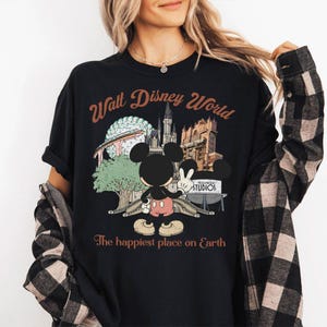 May include: Black t-shirt featuring a graphic of Mickey Mouse in front of Walt Disney World landmarks. The text "Walt Disney World" is arched above the image, with "The happiest place on Earth" below. The shirt has rolled-up sleeves.