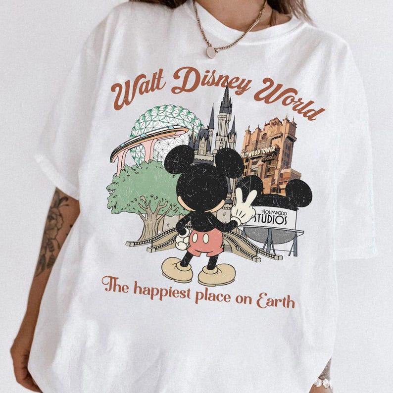 May include: White t-shirt featuring a vintage-style graphic of Mickey Mouse giving a peace sign in front of Walt Disney World landmarks. The text "Walt Disney World" and "The happiest place on Earth" are printed above and below the graphic.