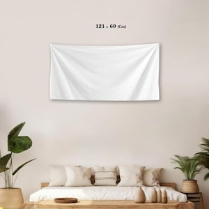 May include: A white rectangular wall tapestry, measuring 121 x 60 cm, hangs on a white wall. The fabric is slightly creased, suggesting a soft material. The tapestry is displayed above a bed with white pillows and a wooden frame.