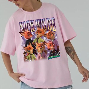 Puede incluir: Camiseta rosa claro con un dise&ntilde;o de Nick Wilde de Zootopia. El dise&ntilde;o incluye el texto "NICK WILDE" sobre un grupo de zorros de dibujos animados. La palabra "ZOOTOPIA" est&aacute; en la parte inferior.