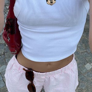 May include: White ribbed tank top with a Minnie Mouse graphic and a gold necklace with a cross pendant. Pink and white striped shorts and brown sunglasses. Red handbag and white sneakers.