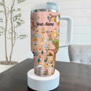 May include: A pink insulated tumbler featuring a whimsical design with various Winnie the Pooh characters. The tumbler has a white handle and lid, with the text "Your Name" displayed prominently. The design includes illustrations of Pooh, Tigger, and other characters.