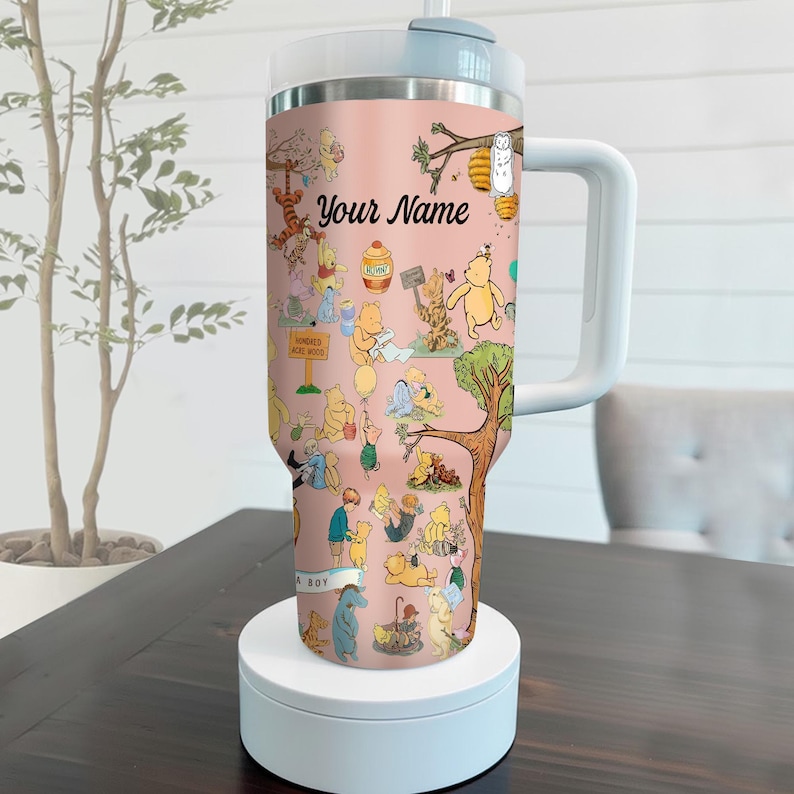 May include: A pink insulated tumbler featuring a whimsical design with various Winnie the Pooh characters. The tumbler has a white handle and lid, with the text "Your Name" displayed prominently. The design includes illustrations of Pooh, Tigger, and other characters.