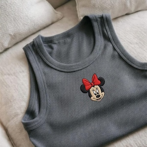 May include: Gray ribbed tank top featuring an embroidered Minnie Mouse design with a red bow. The sleeveless top is made of a soft material and is suitable for casual wear. The top is laying on a light-colored surface.
