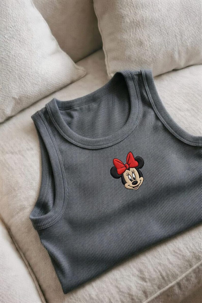 May include: Gray ribbed tank top featuring an embroidered Minnie Mouse design with a red bow. The sleeveless top is made of a soft material and is suitable for casual wear. The top is laying on a light-colored surface.