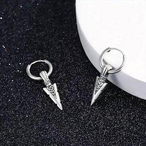 May include: Two silver-colored earrings with a Celtic knot design on a black background. Each earring features a hoop and a pendant shaped like an arrowhead. The earrings are made of a shiny metal.