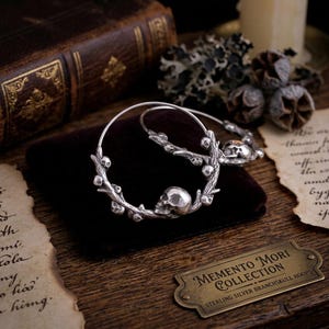 Puede incluir: Un par de pendientes de aro de plata de ley de la colección Memento Mori. Cada pendiente presenta un diseño de calavera y rama. Los pendientes se exhiben sobre una superficie de terciopelo burdeos oscuro, con un libro antiguo y una placa de latón.