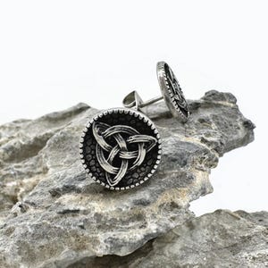 May include: Silver-toned stud earrings featuring a Celtic knot design. The earrings have a circular shape with a textured background and a beaded border. One earring is in focus, while the other is slightly behind it.