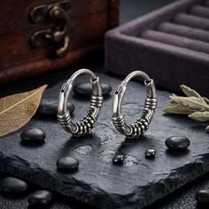 May include: Two silver hoop earrings with a tribal design. The earrings have a thick band with a series of etched lines and a braided pattern. The earrings are displayed on a black surface with small black stones.