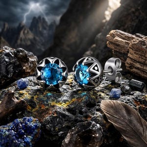 May include: Two silver stud earrings with a blue gemstone center. The earrings have a decorative, openwork design around the gemstone. The image is set against a rocky, mountainous background with a stormy sky.