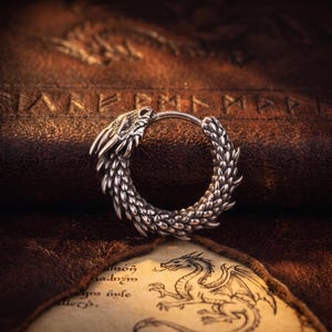 May include: A silver dragon-shaped earring. The earring is a hoop, with the dragon's head at one end and a scaled body forming the hoop. The background is a textured brown surface with a parchment-like paper.
