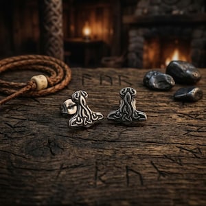 Pode incluir: Um par de brincos prateados em forma de martelo de Thor, Mjolnir, com intrincados desenhos de nós. Os brincos são exibidos numa superfície de madeira envelhecida, evocando uma estética rústica de inspiração viking.