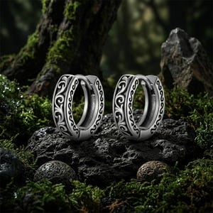 Peut inclure: Deux boucles d'oreilles créoles argentées avec un motif tourbillonnant orné. Les boucles d'oreilles sont présentées sur une roche sombre et texturée, entourée de mousse et de feuillage verts. Le design est détaillé et symétrique.