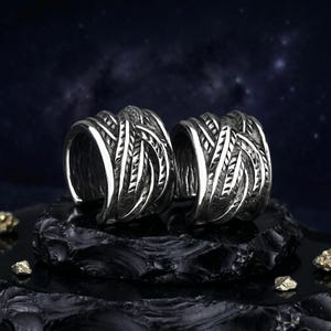 May include: Two silver-colored rings with a woven design. The rings are displayed on a black, textured surface against a dark blue background with a starry effect. The rings have a thick band with detailed, textured patterns.