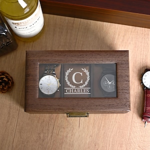 May include: A rectangular dark brown wooden watch box with a glass lid featuring the engraved letter C inside a laurel wreath and the name CHARLES. The box contains two wristwatches, one with a silver face and one with a black face.