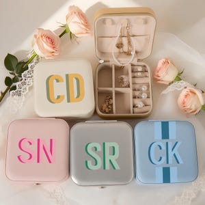 May include: A selection of personalised square jewellery boxes in cream, pink, silver, and blue. Each box has initials in contrasting colours. One box is open, revealing jewellery. Roses are arranged around the boxes.