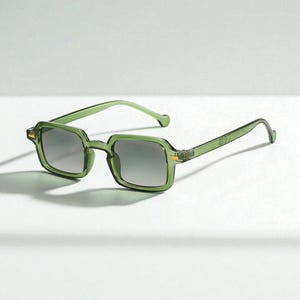 Op de afbeelding: Groene zonnebril met vierkant montuur. De zonnebril heeft een doorschijnend groen montuur en verloopglazen. De pootjes zijn dun en buigen naar de oren. De zonnebril staat tegen een witte achtergrond.