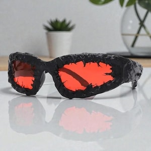 Può includere: Occhiali da sole neri con lenti rosse. Le montature hanno un aspetto strutturato e ruvido. Le lenti sono di un rosso brillante e vibrante. Gli occhiali da sole sono appoggiati su una superficie bianca, con una piccola pianta sullo sfondo.