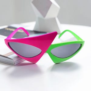 Puede incluir: Gafas de sol inusuales en rosa y verde brillante. Las monturas tienen un diseño angular, con un lado rosa y el otro verde. Las lentes son de color gris oscuro. Un accesorio de moda audaz.